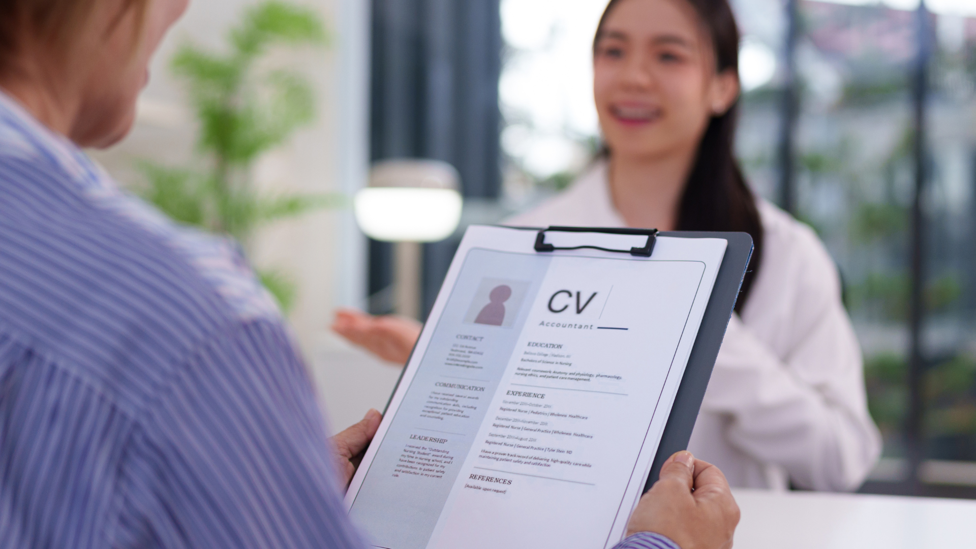 Person reviewing a CV during a job interview with a smiling candidate in the background.