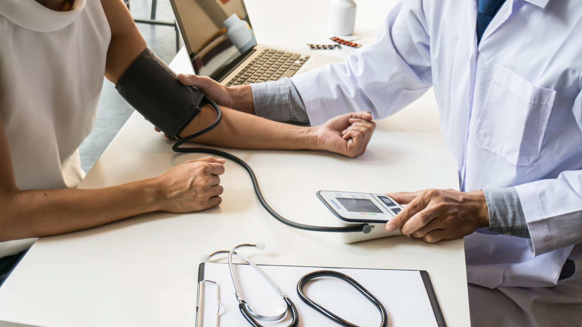 Doctor taking patient's blood pressure in a clinic. White coat, medical equipment, and desk visible.