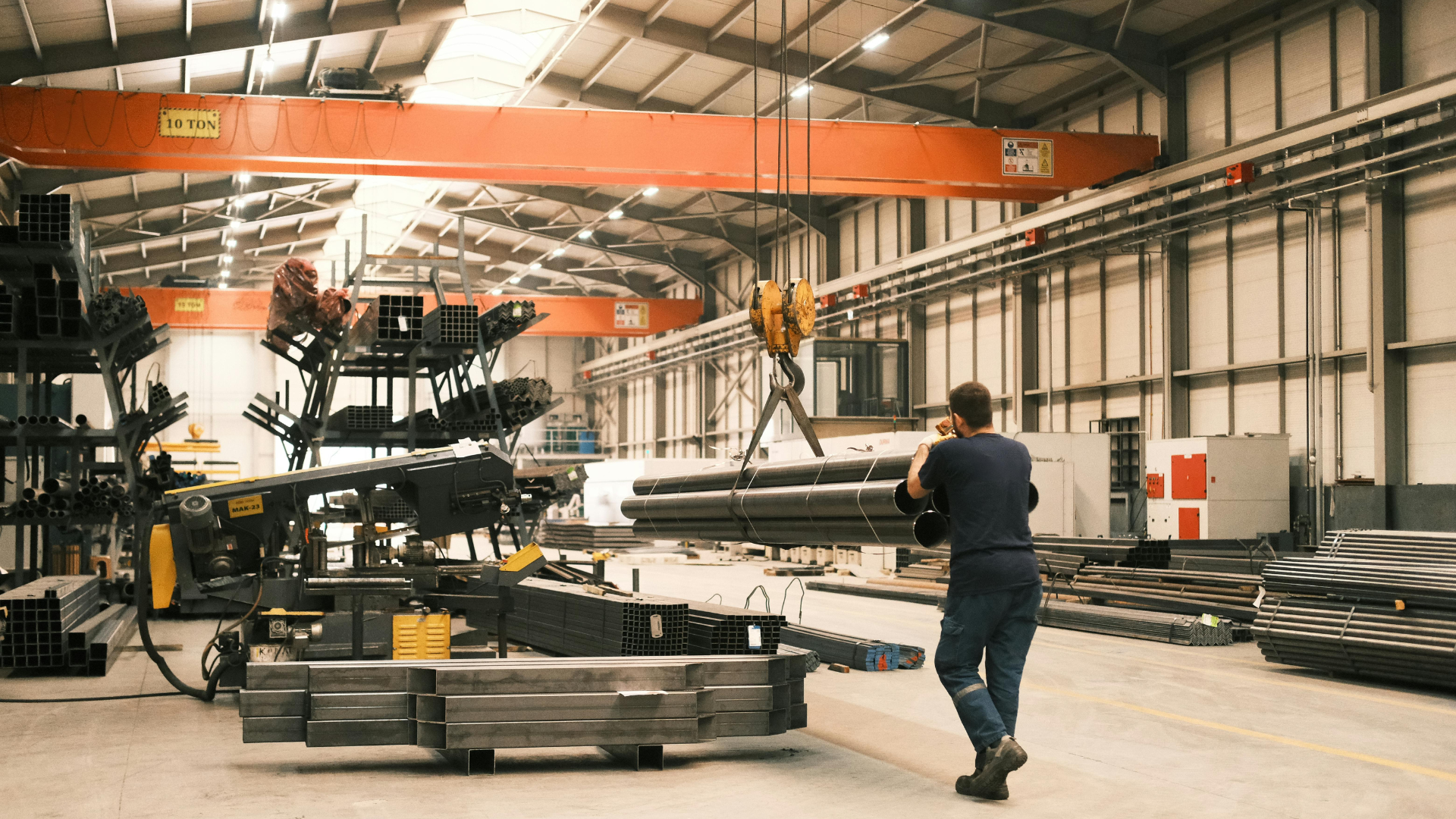 Man in metal factory, watching overhead crane lifting metal pipes. Warehouse setting, natural light.