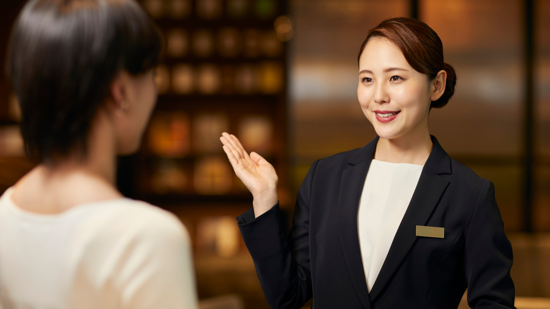 Hotel receptionist smiling, gesturing toward a guest; indoor setting.