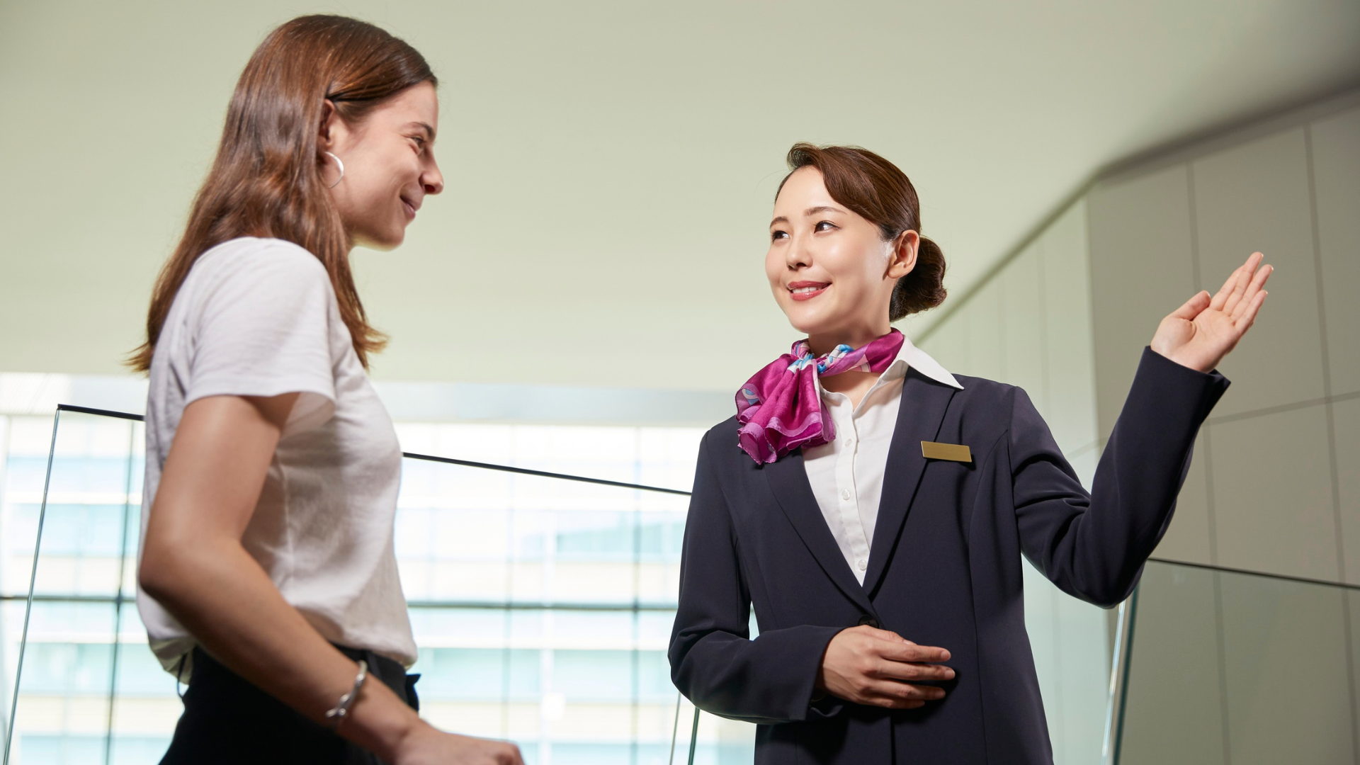 Woman in white shirt speaking to a uniformed attendant pointing right in a bright hallway.