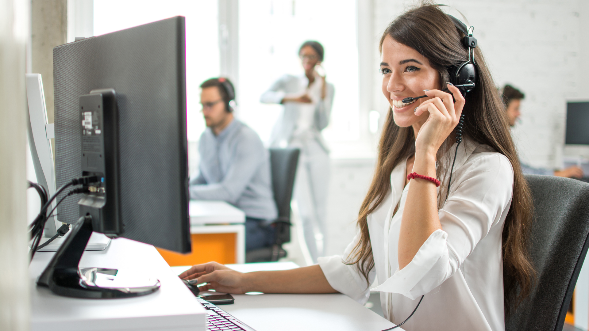 Woman with headset smiles while working at a computer in an office setting.