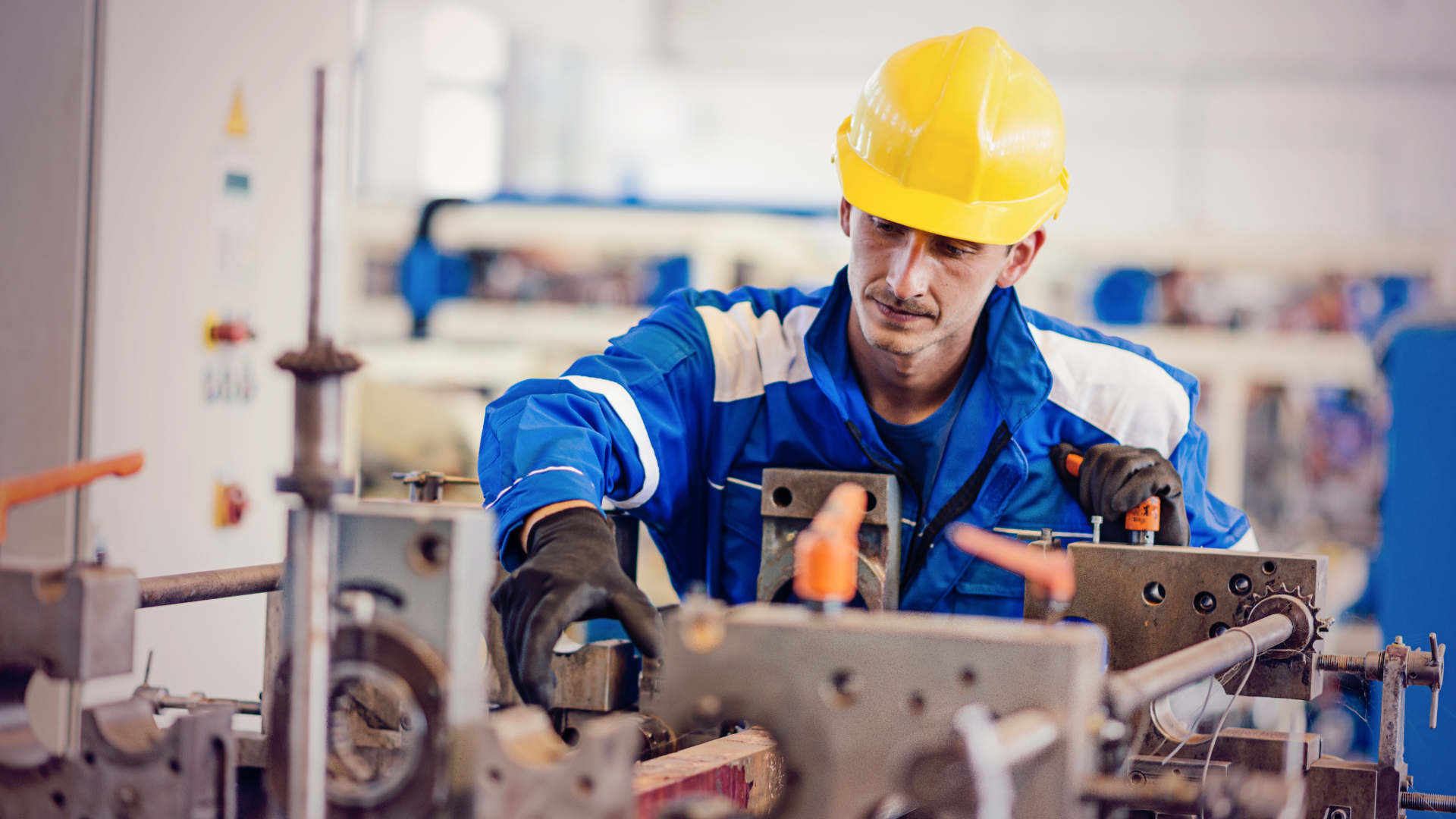 Man in yellow hard hat and blue coveralls working on machinery in a factory.