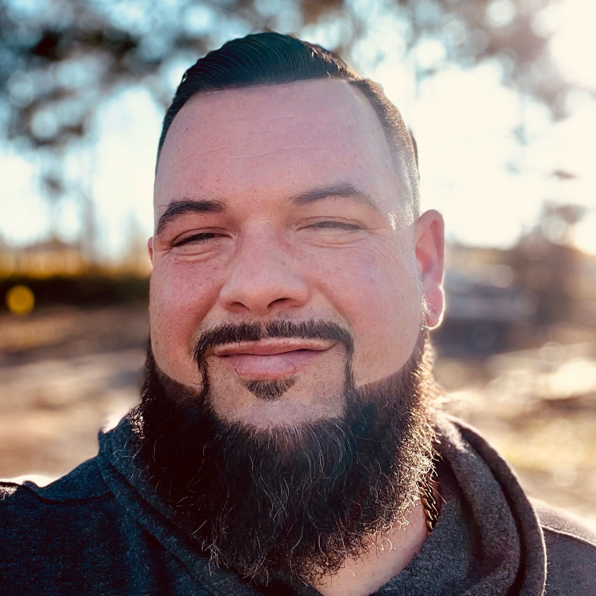 A man with a beard and a gold chain is smiling in front of a field of leaves.