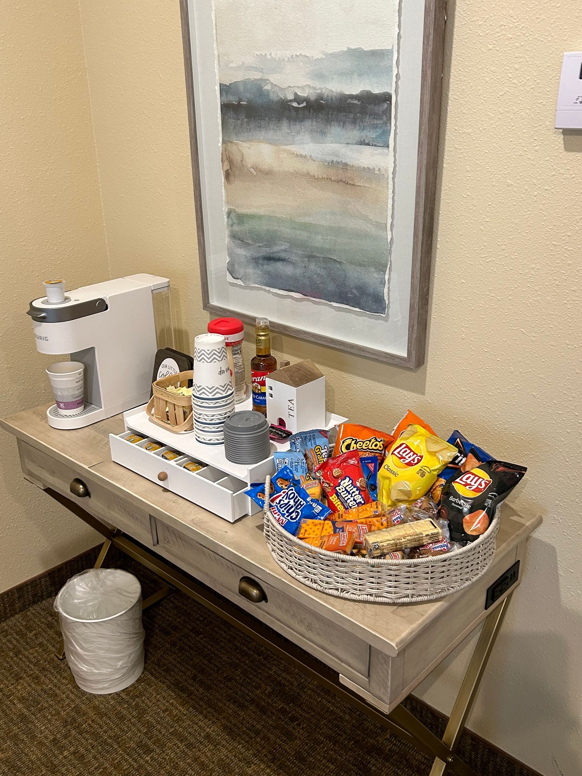 There is a coffee maker and a basket of snacks on the table.