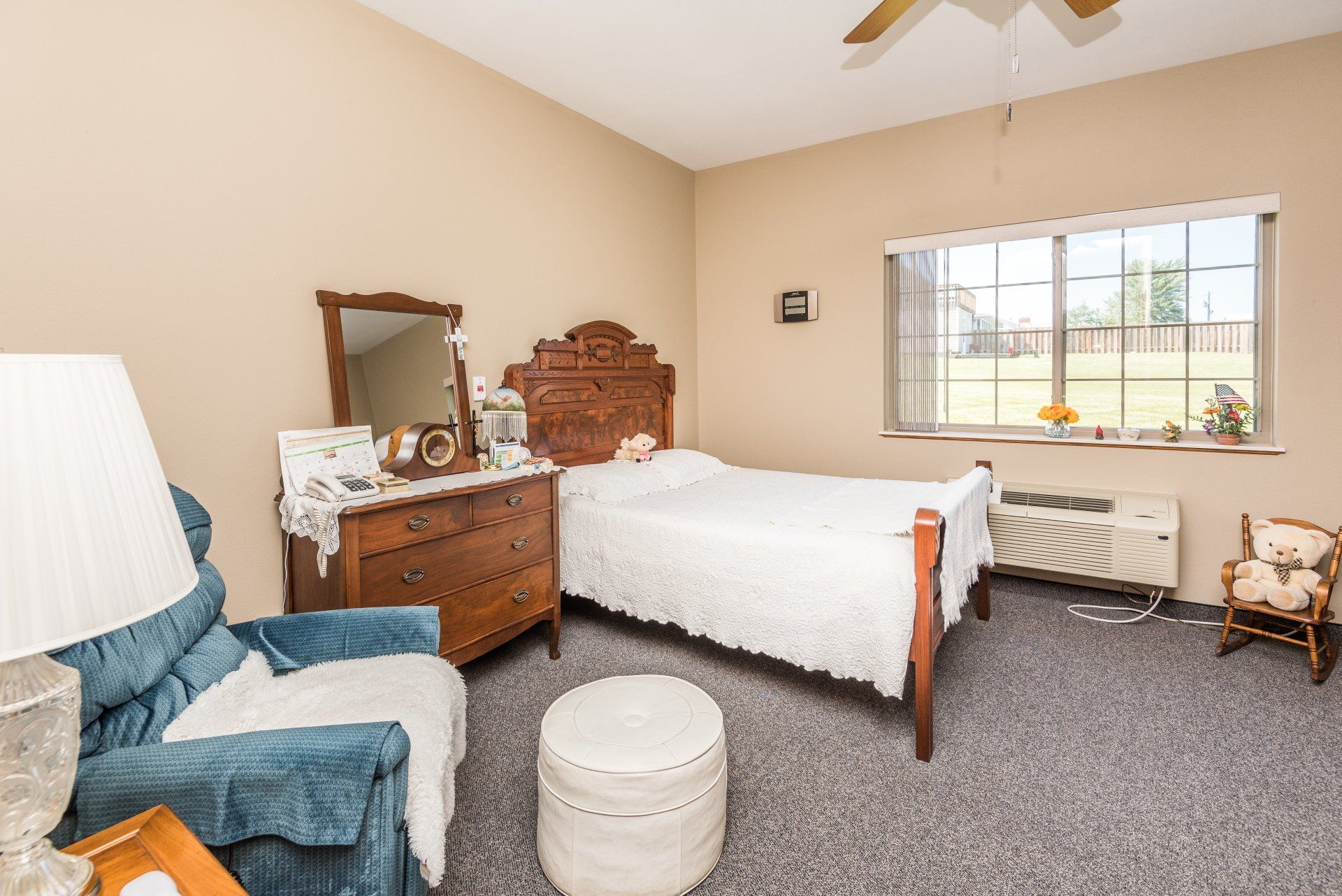 A bedroom with a bed , dresser , chair , lamp and window.