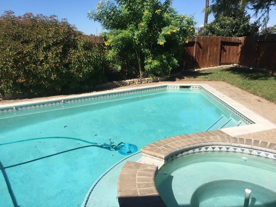 A backyard pool with turquoise water, a hot tub, and surrounding landscaping under a sunny sky.