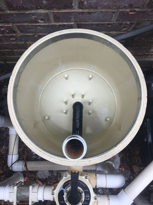 Overhead view of an open, beige swimming pool filter housing with a black pipe in the center and PVC pipes below.