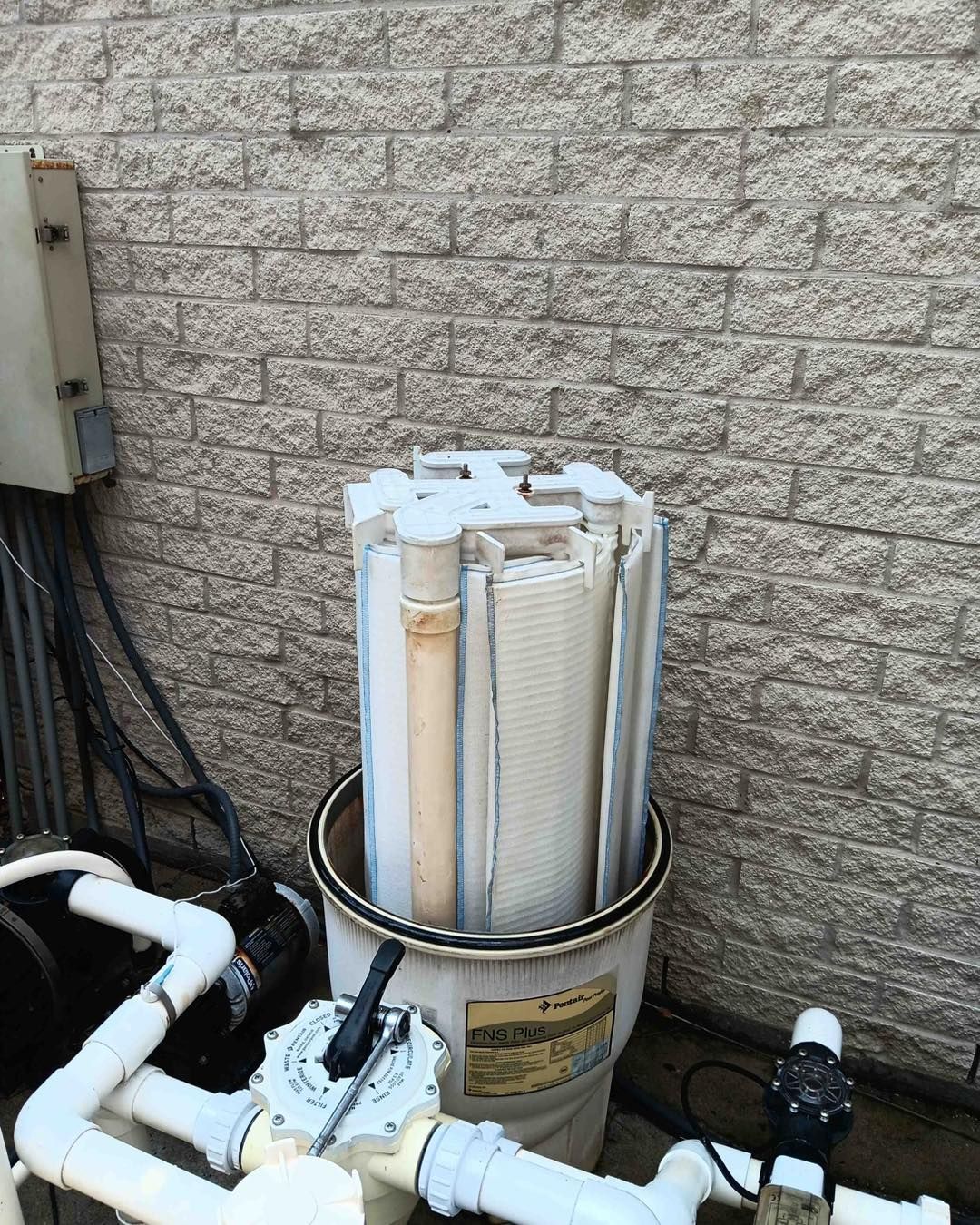 A white pool filter cartridge in a bucket next to pool plumbing and a brick wall.