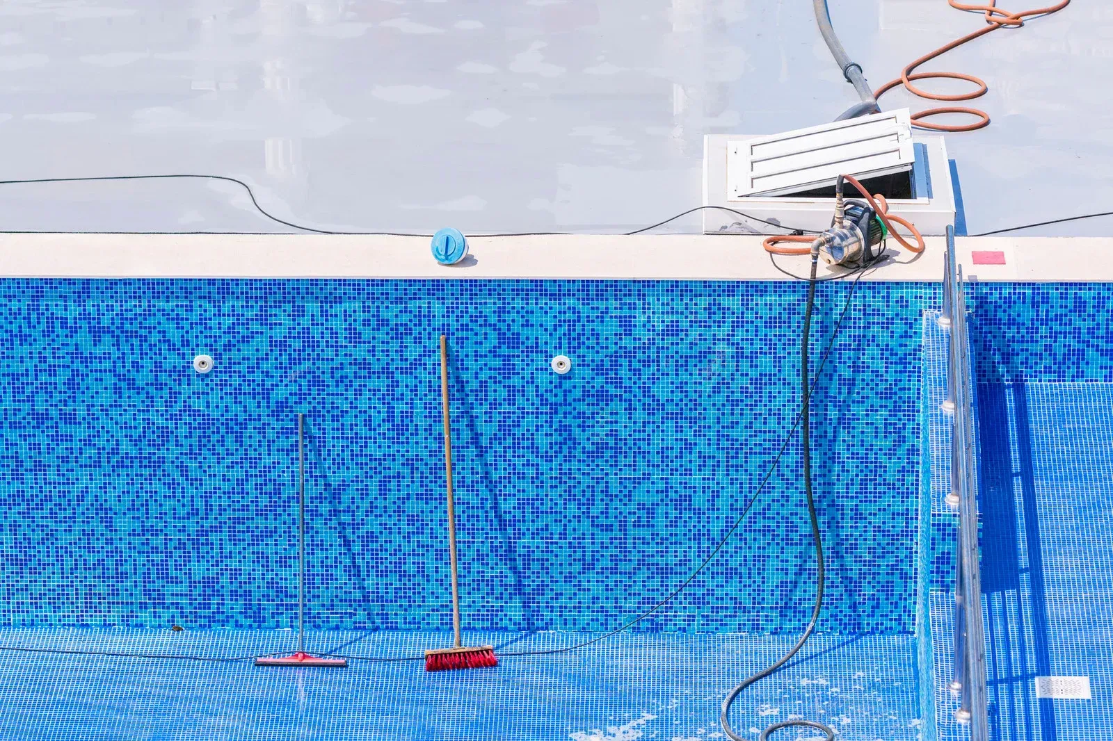 Empty blue-tiled swimming pool with cleaning tools, electrical equipment, and hose on the edge.