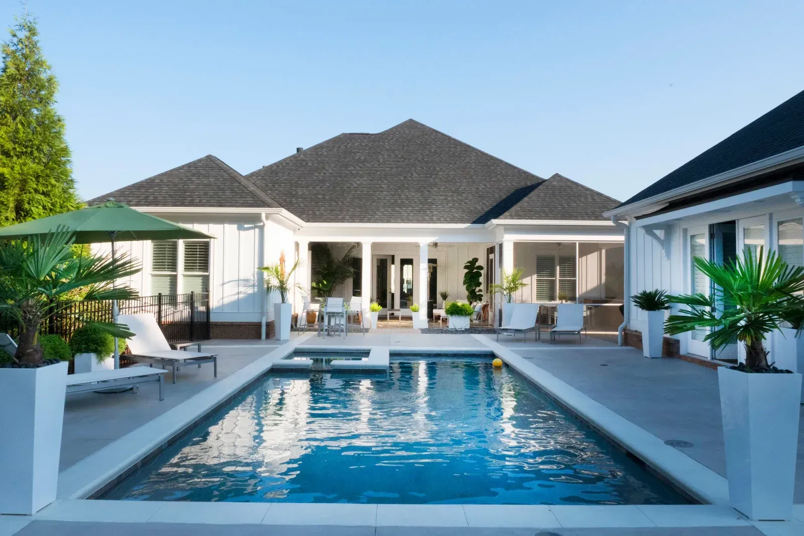 Backyard with a rectangular pool, white patio furniture, and a white house under a clear blue sky.