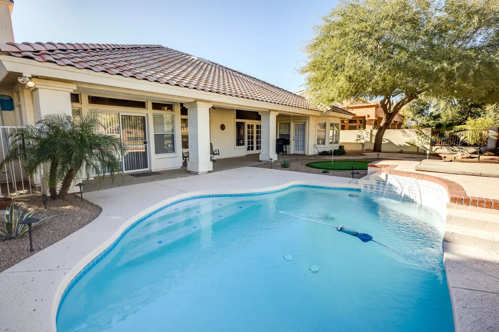 Backyard with a blue swimming pool, white house, and a mature tree under a bright sky.