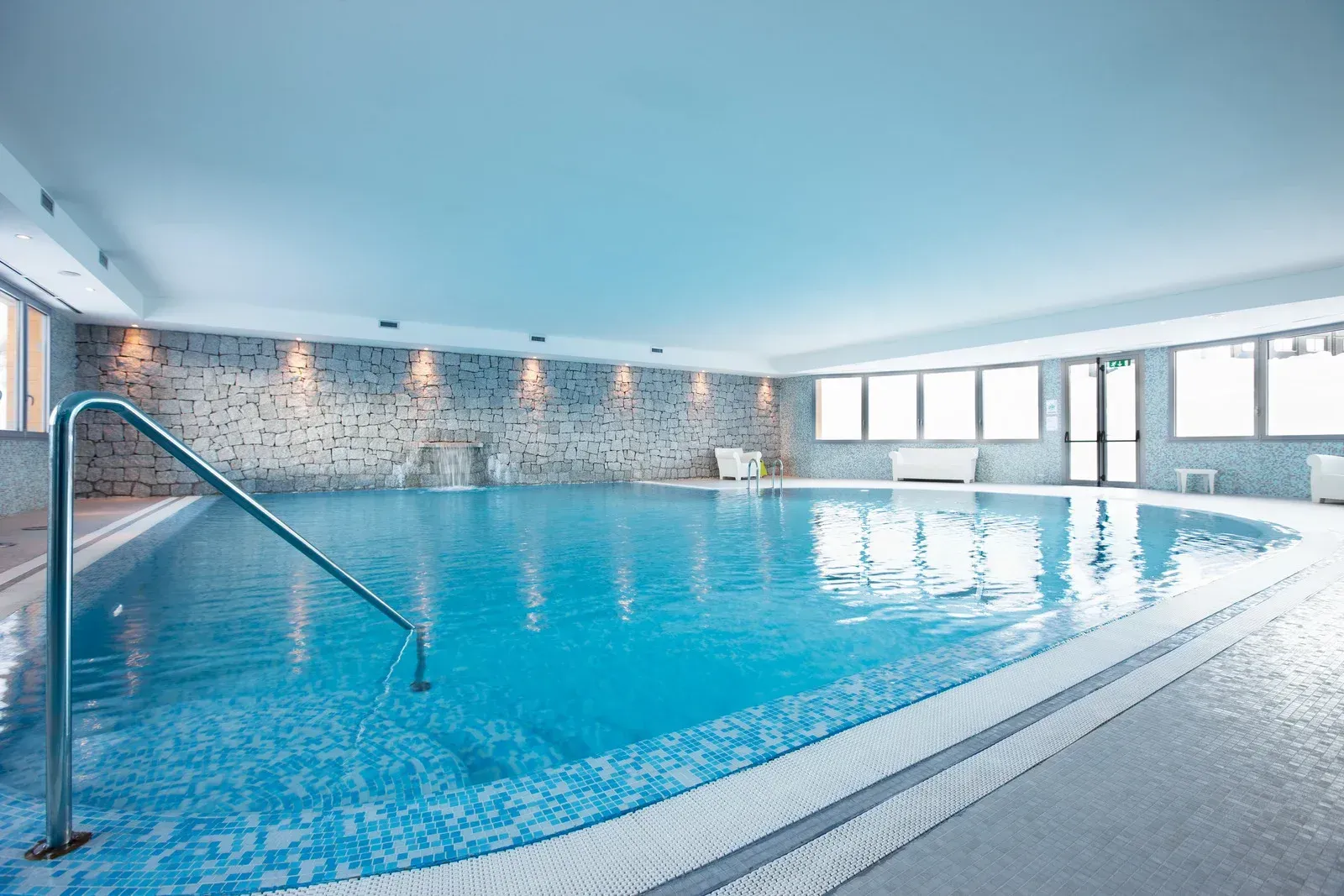 Indoor swimming pool with blue tiled walls and water, handrail on the side, and large windows.