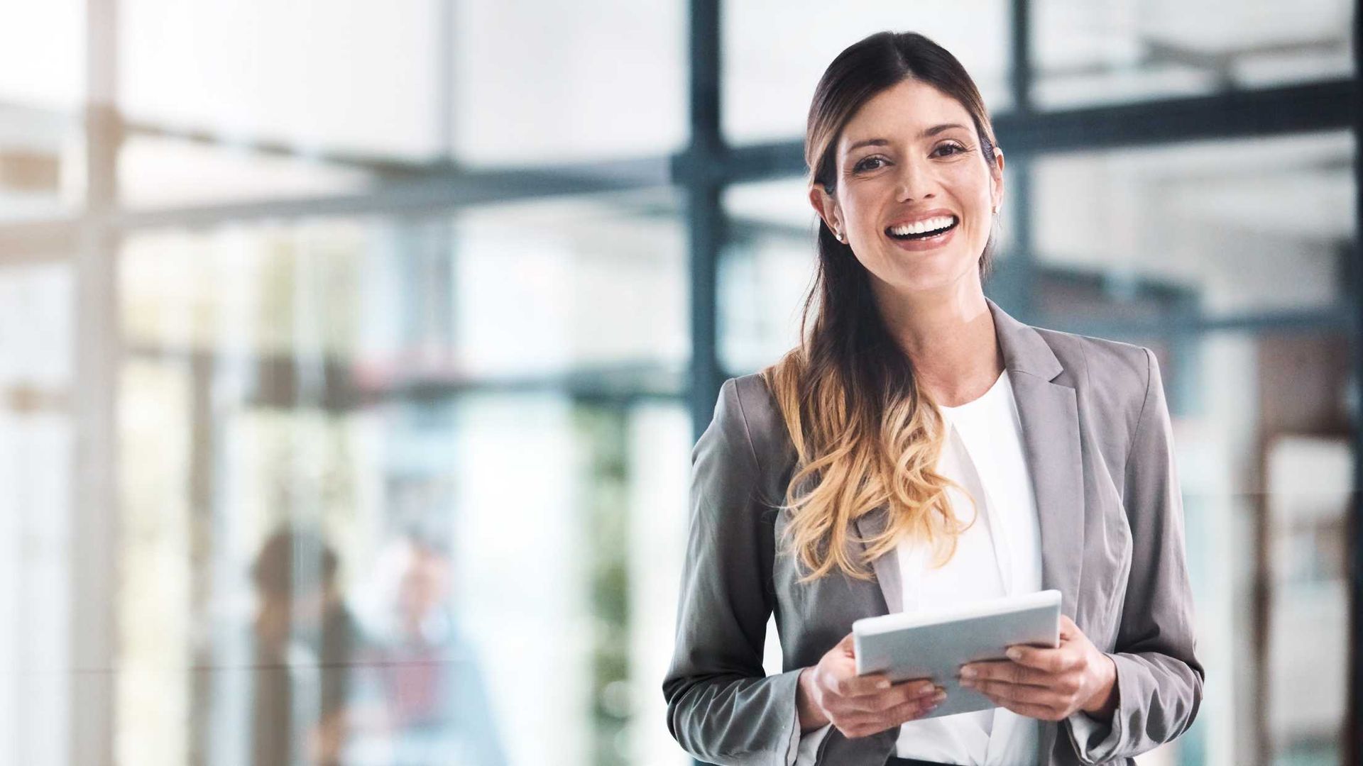 Woman in a gray suit smiles while holding a tablet in a bright office setting.