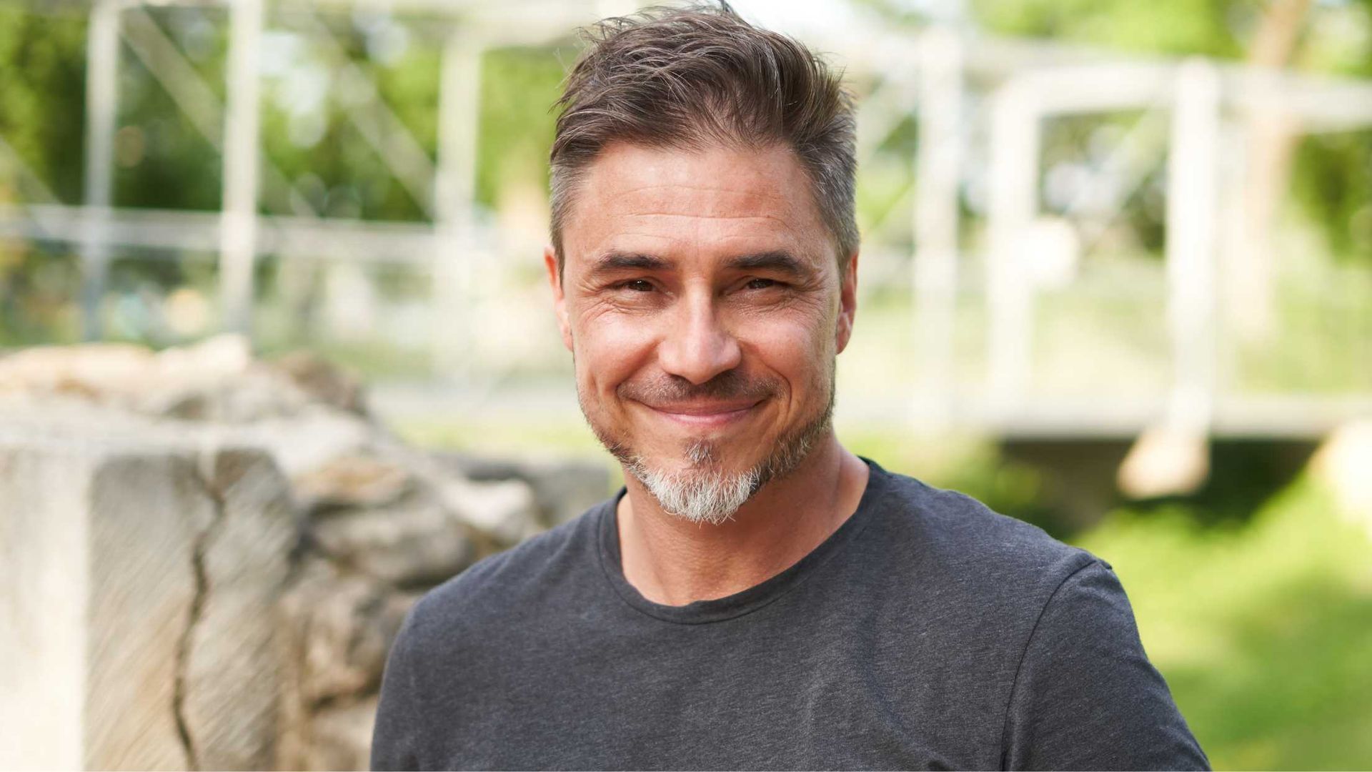 Man with graying hair and beard smiles outdoors.
