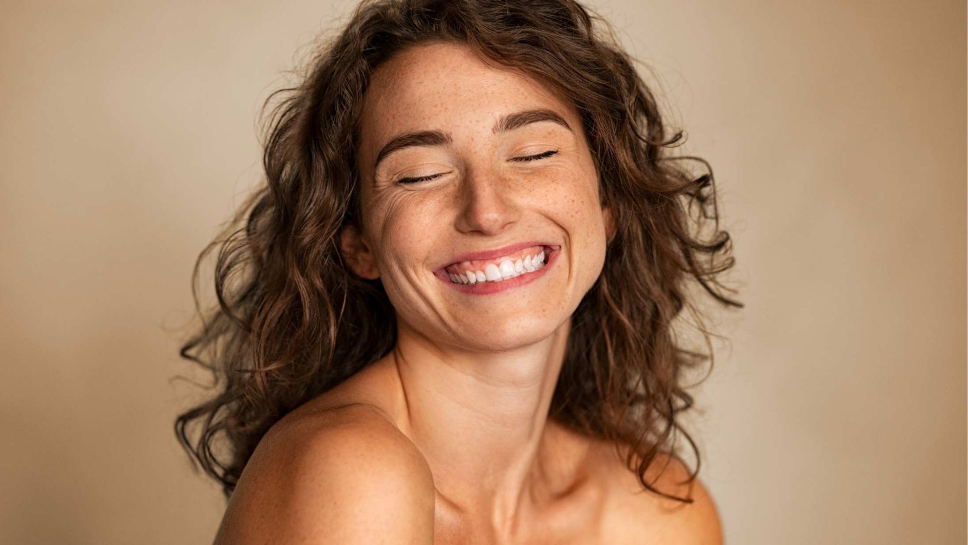 Woman with freckles and curly brown hair smiles broadly with closed eyes.