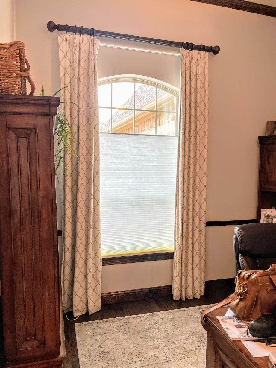Window with arched top, framed by patterned drapes and cellular shades. Wooden furniture, rug, and natural light in the room.