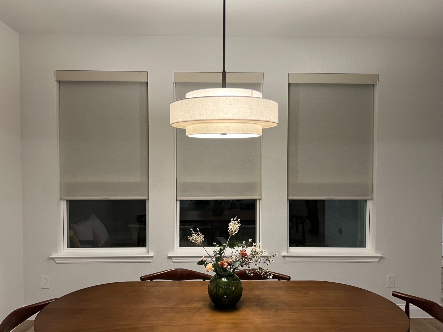 Dining room with a round wooden table, three windows with shades, and a layered pendant light.