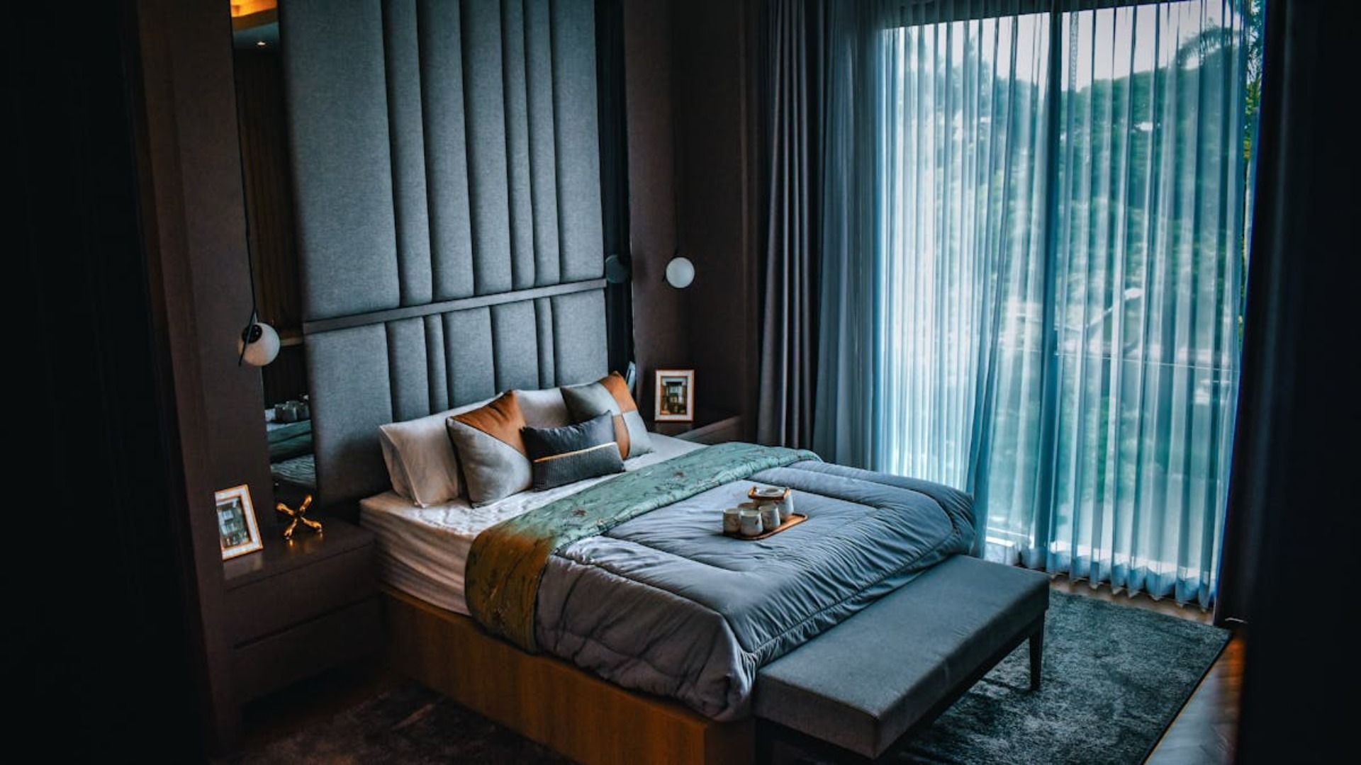 Dormitorio con gran cabecero tapizado, cama con ropa de cama gris y cortinas transparentes.