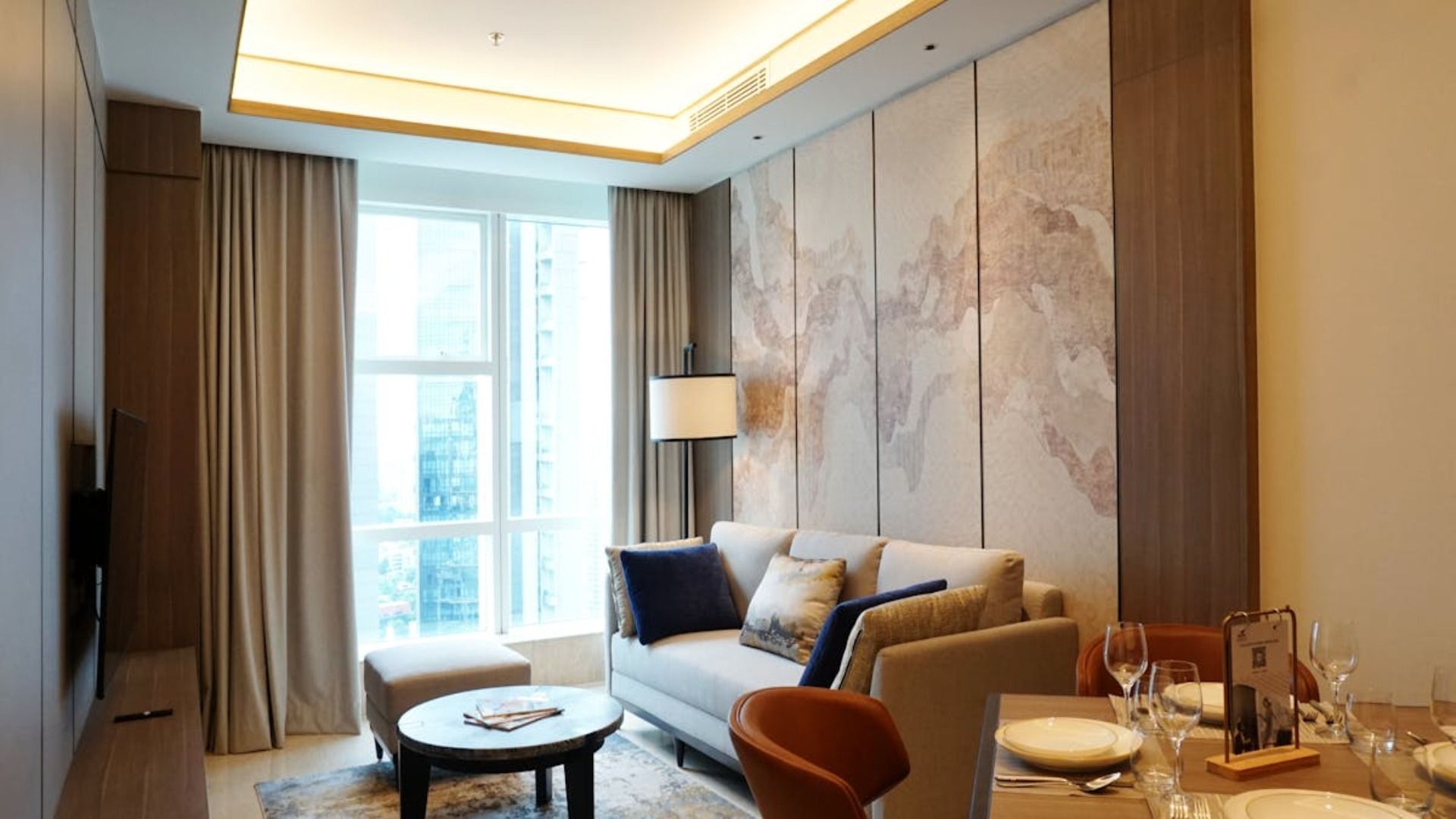 Sala de estar con sofá, mesa de centro y ventanal. Tonos neutros, paneles decorativos y mesa de comedor.