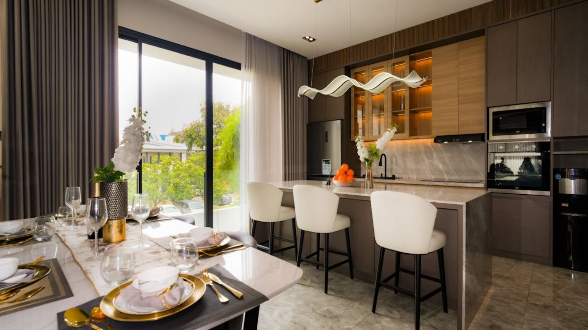 Moderna cocina y comedor con isla, taburetes y mesa preparada para comer, gran ventanal con vista al exterior.