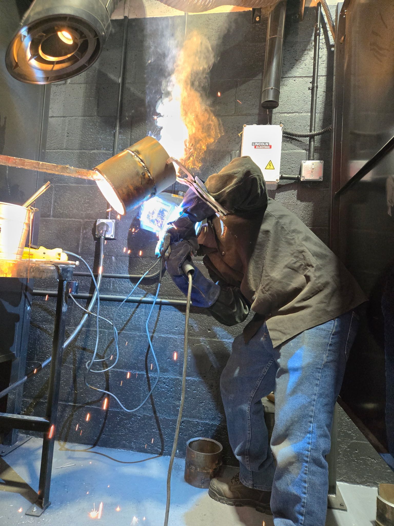 A person in protective gear uses a welding torch on a pipe inside an industrial workshop, emitting sparks and bright light.
