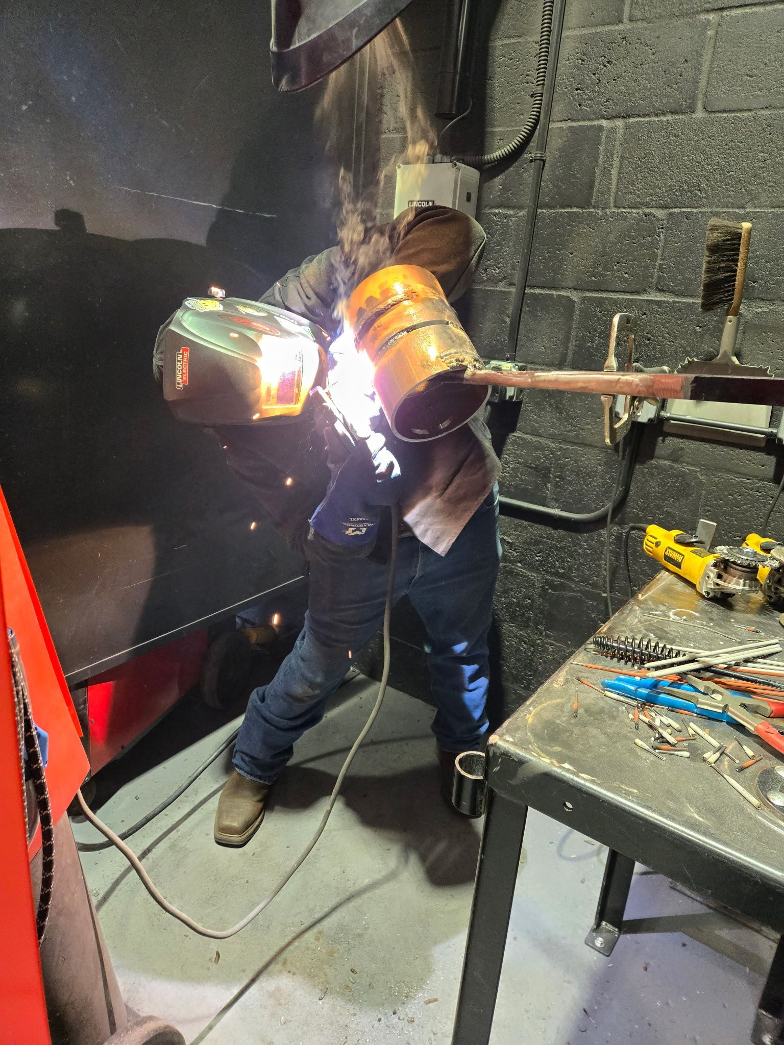 A person wearing a welding helmet and protective gear arc welds metal in a workshop, sparks flying at the connection.