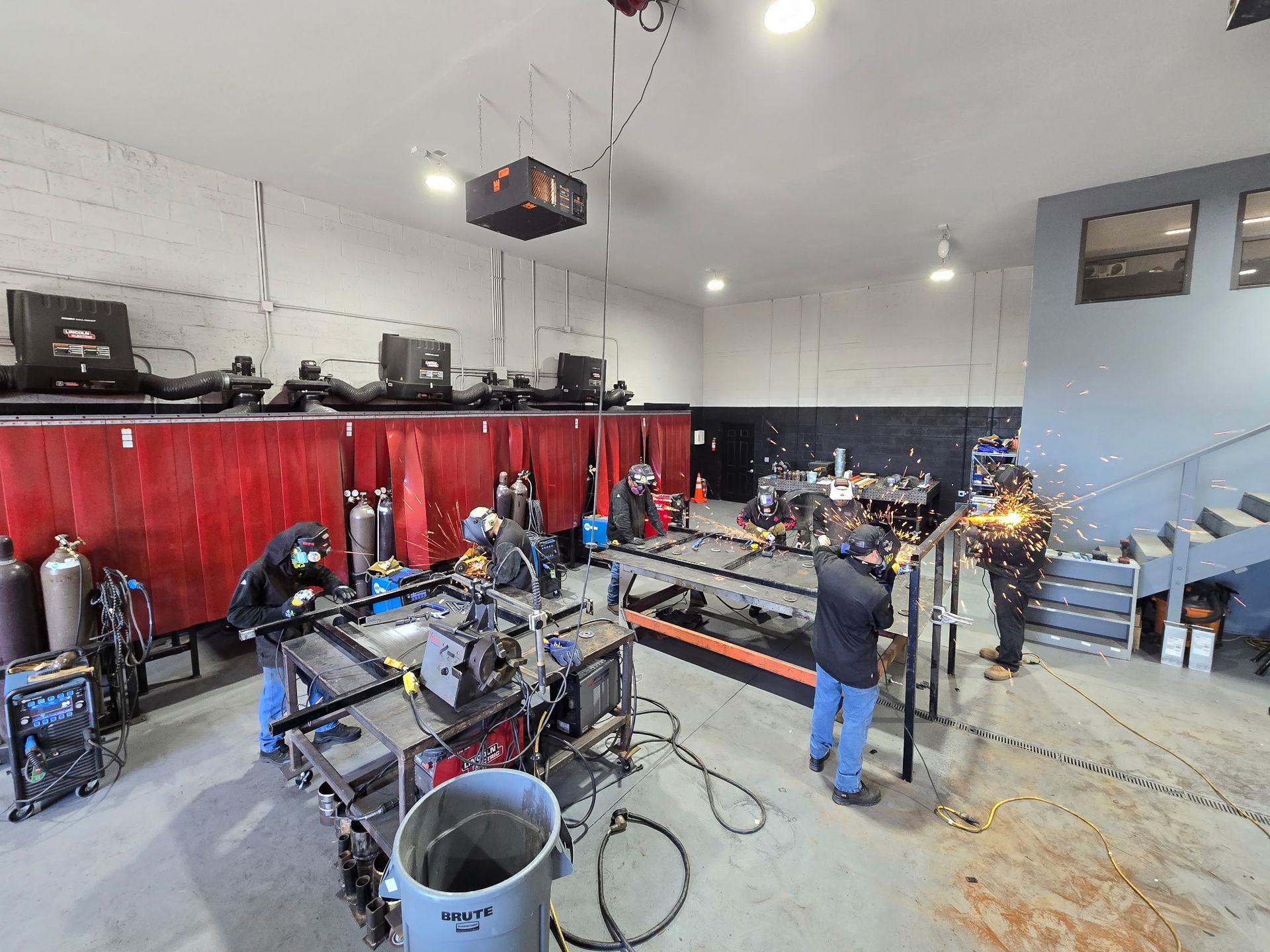 Welders work in a well-lit shop with red privacy curtains, welding equipment, and sparks flying in the background.