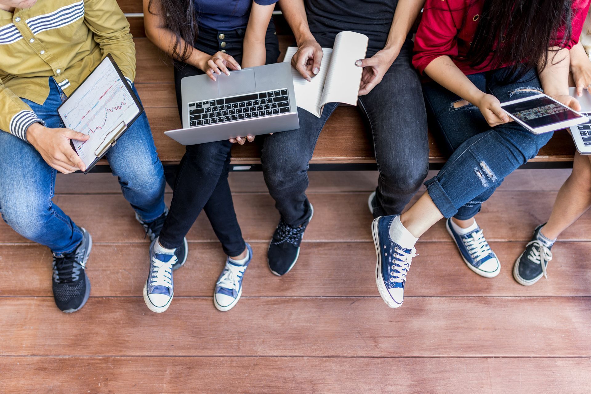 Five people sitting on a wooden bench with laptops, notebooks, and tablets, with their legs in view.