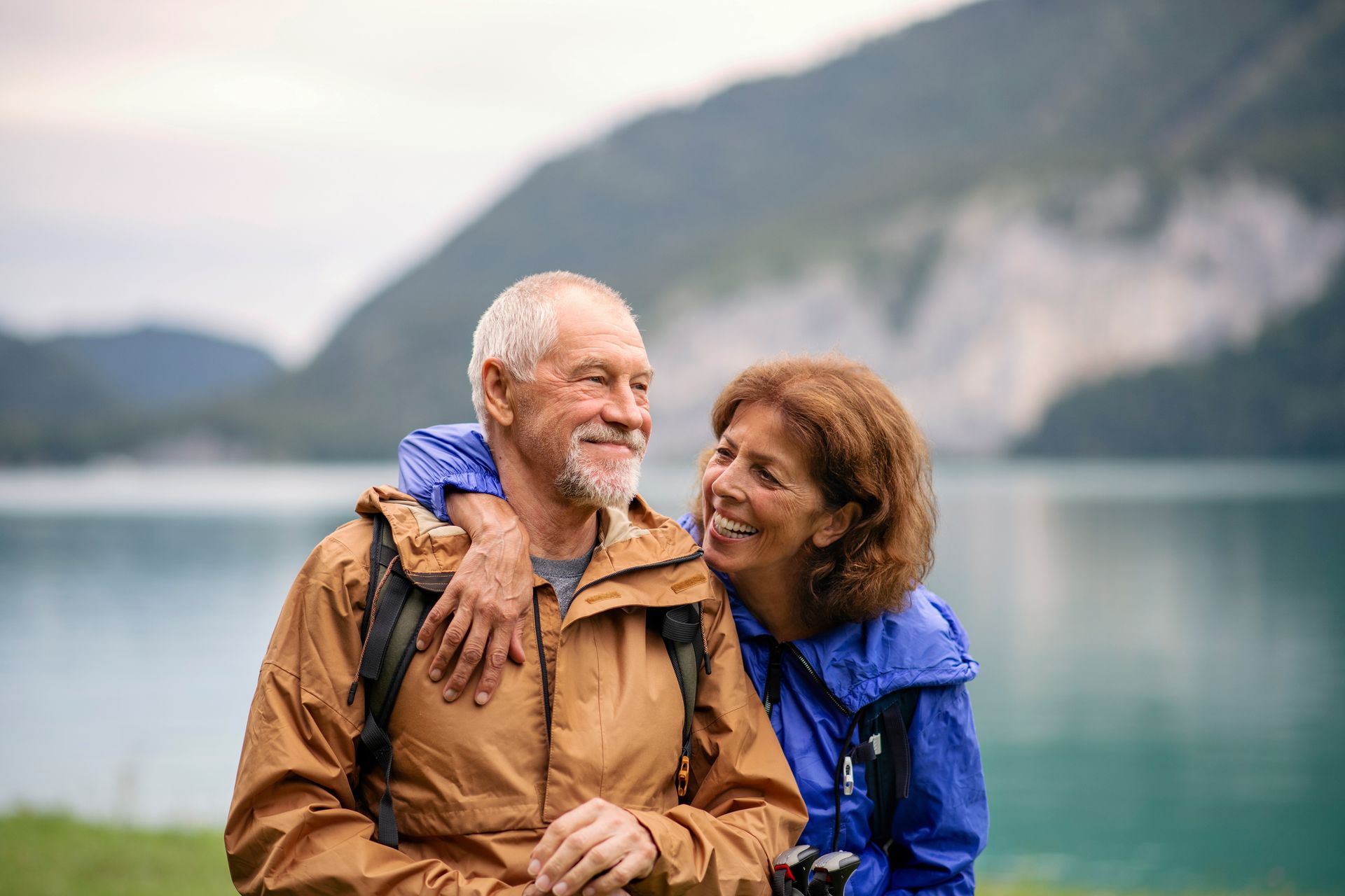 Elderly couple posing happily in front of a scenic lake with mountains in the background.