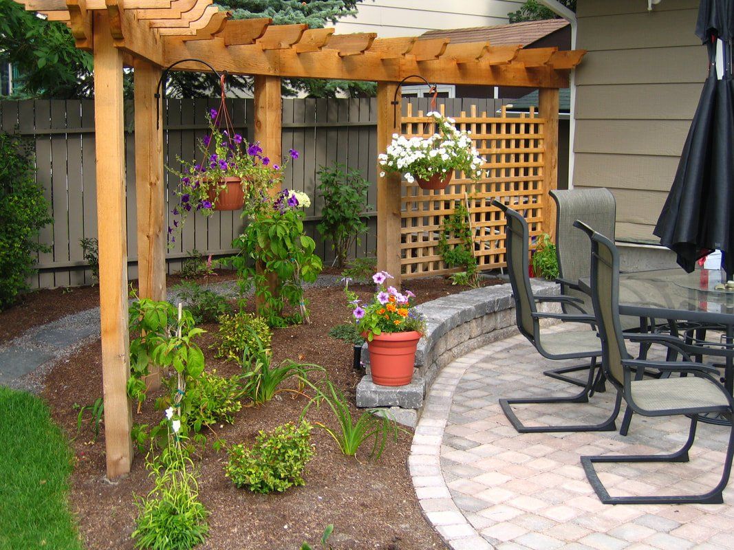 Trellis with hanging plants over garden growing vegetables behind paved circular patio with lawn chairs