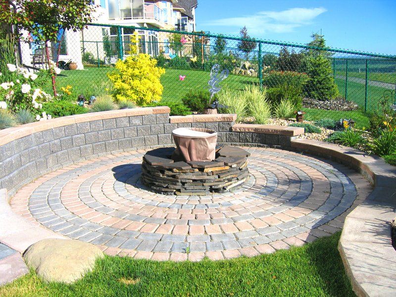 Custom backyard landscape paved patio with an outdoor fireplace surrounded by bushes and a grass lawn