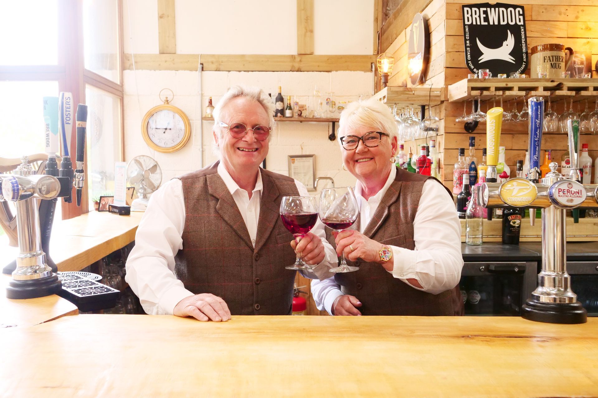 A man and a woman are standing behind a bar holding wine glasses.
