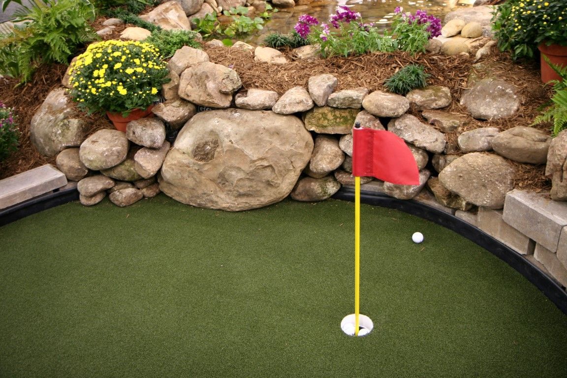 A white golf ball sits on green artificial turf near a hole with a red flag, set against a background of rocks and flowers.
