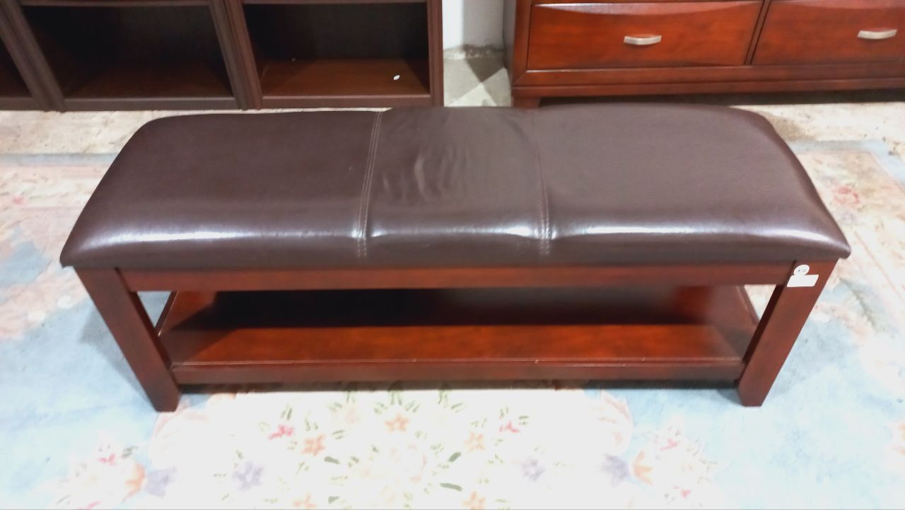 Banco marrón con asiento acolchado y estante inferior, delante de un mueble de madera.