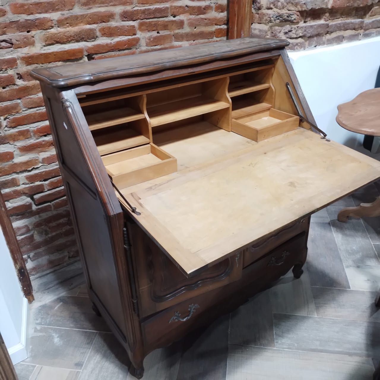 Escritorio de madera con tapa en ángulo, dos puertas y un cajón, contra una pared de ladrillos.