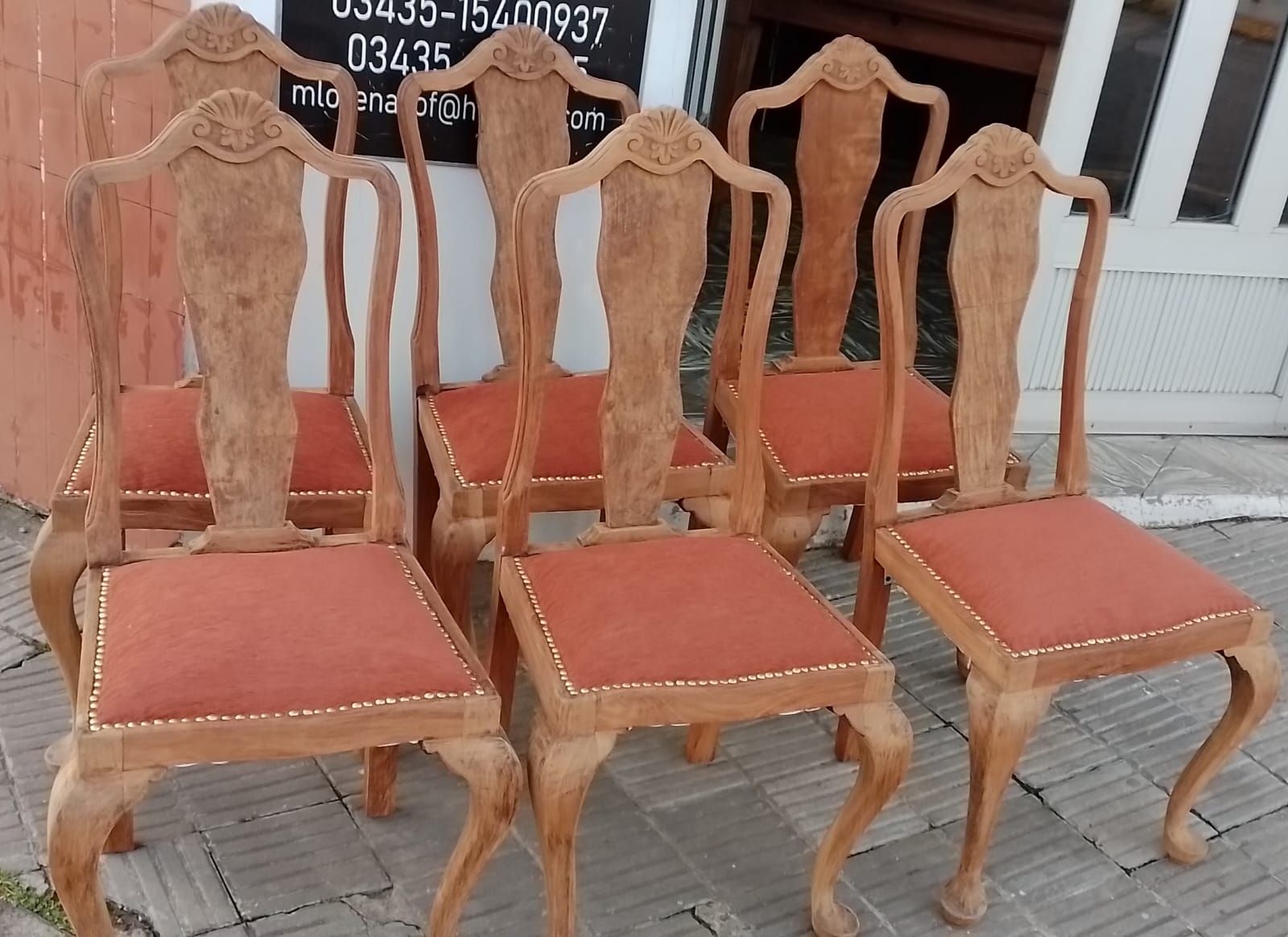 Seis sillas de madera con respaldos ornamentados y asientos tapizados en rojo se exhiben afuera de una tienda.