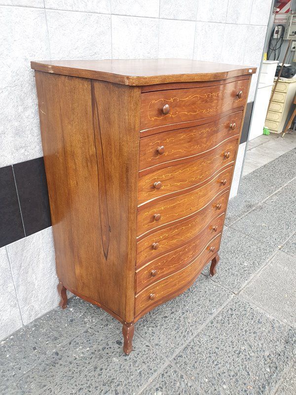 Cómoda antigua de madera con cajones curvos y patas pequeñas contra una pared de azulejos.
