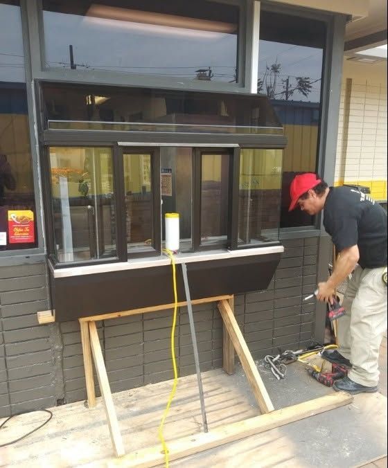 Man in red cap works on a drive-thru window. Window supported by wood frame, cords run through it.