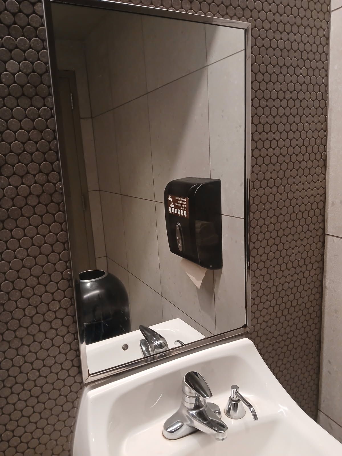 Bathroom sink with mirror, soap dispenser, and paper towel dispenser. Tile wall.