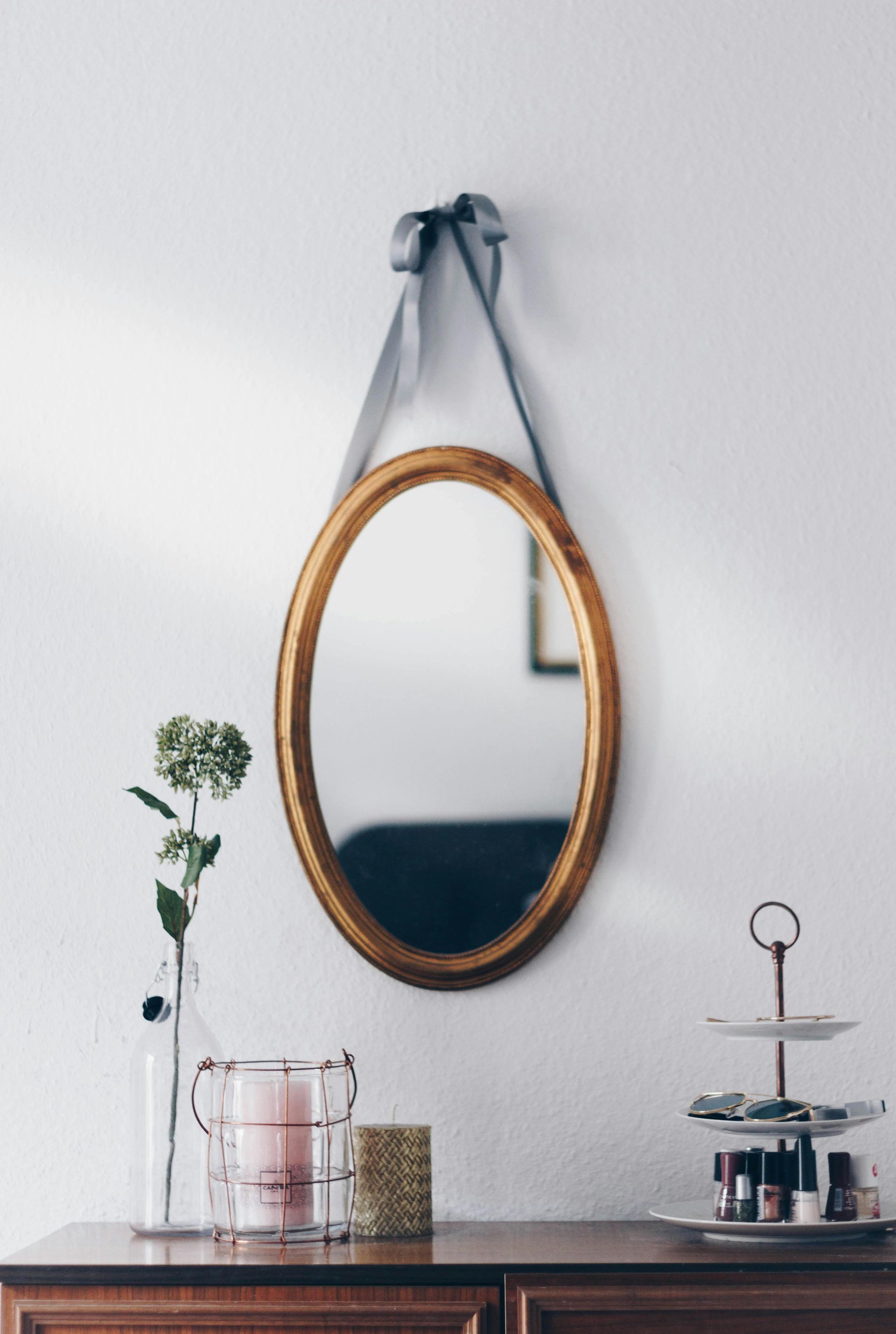 Oval gold-framed mirror hanging on a white wall, over a wooden dresser with decorative items.