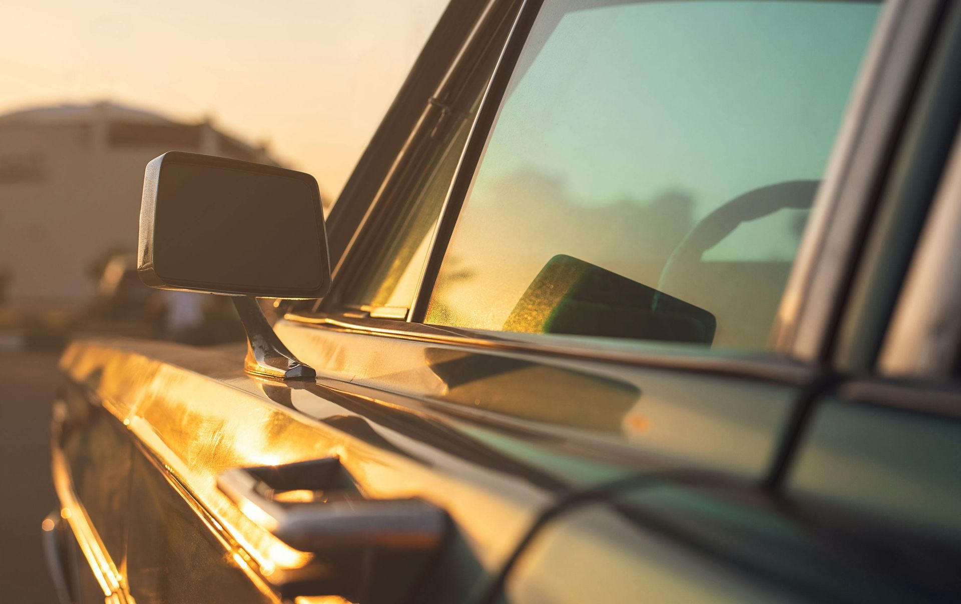 Close-up of a dark-colored car's side, reflecting golden sunlight. The side mirror and window are visible.