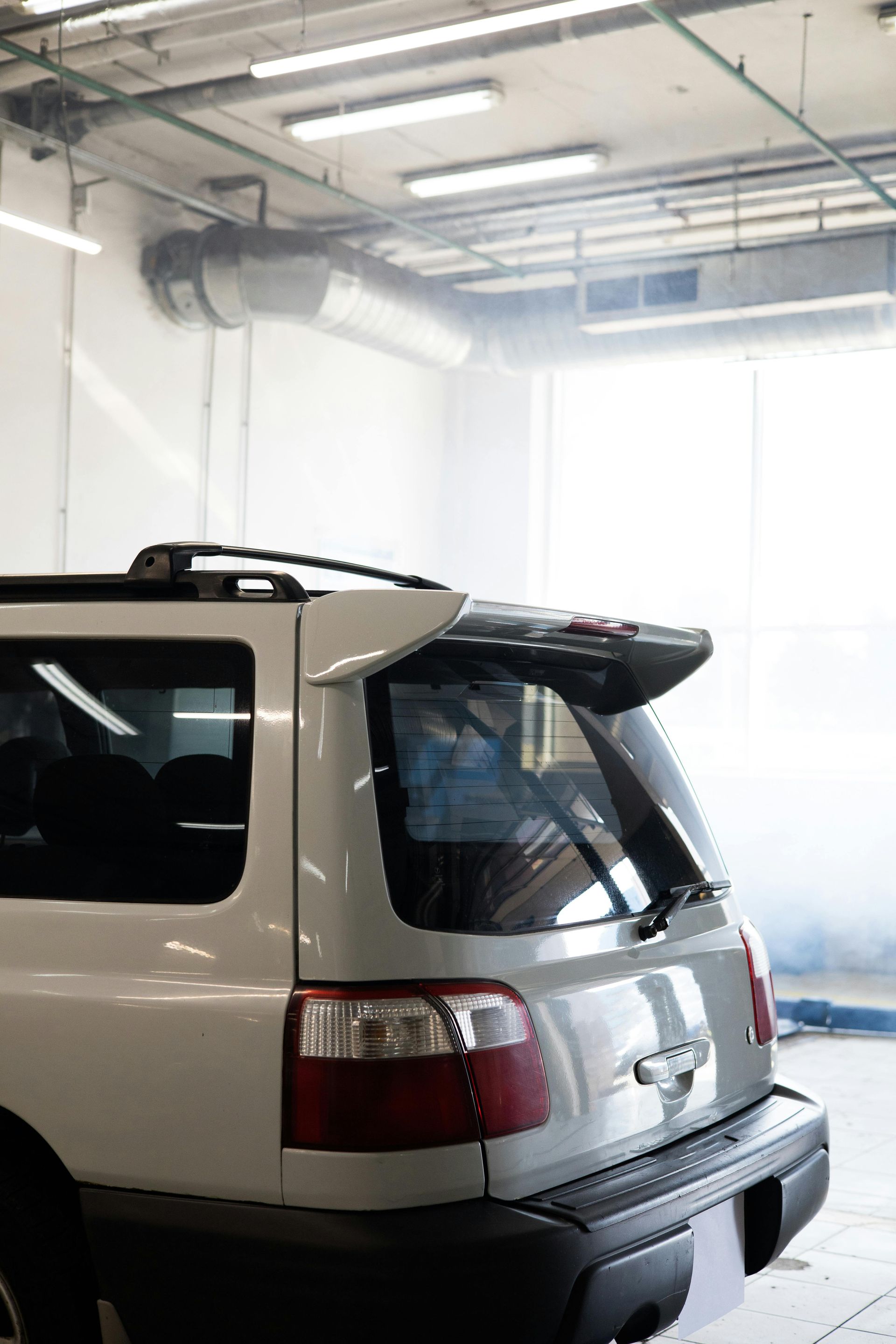 White SUV in a car wash bay, rear view, water vapor, bright lighting.