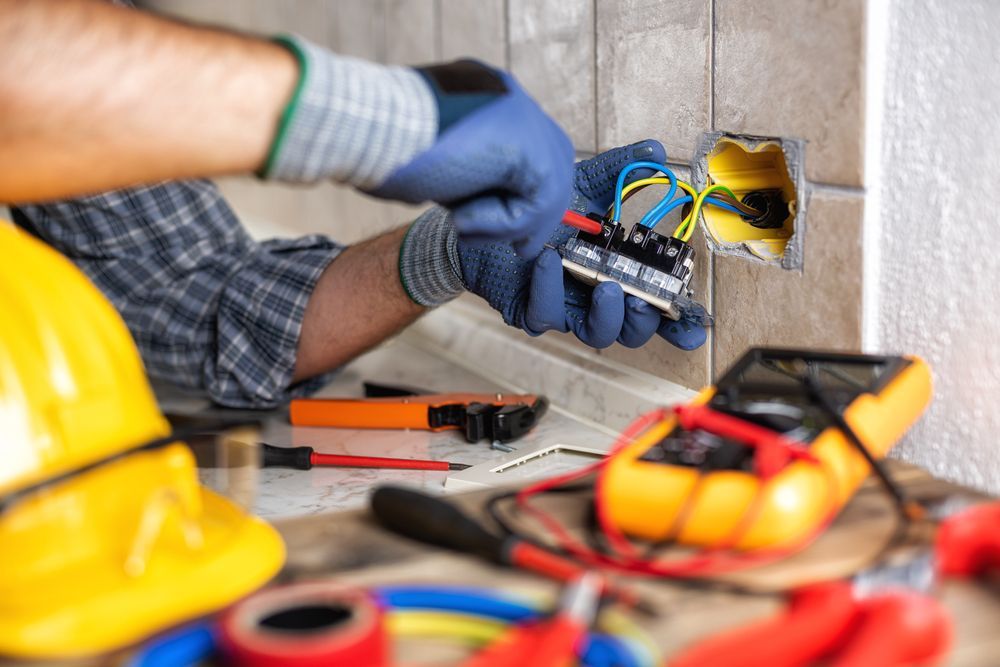 Electrician installing outlet, wearing gloves, using tools, working near a yellow hard hat and multimeter.