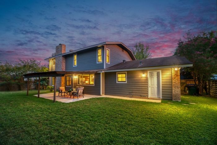 Back of blue house at dusk with patio, green lawn, and colorful sky.