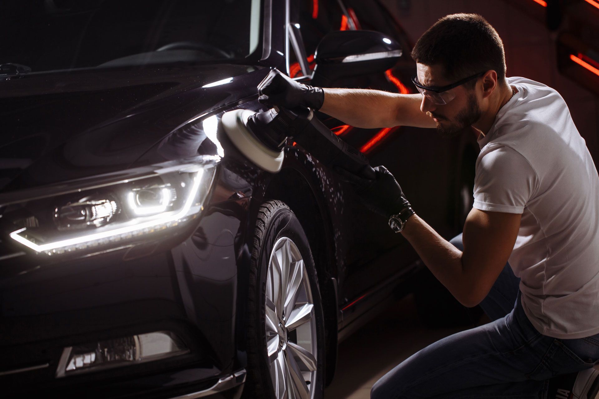 Man polishing a black car with a buffer, wearing gloves and safety glasses.