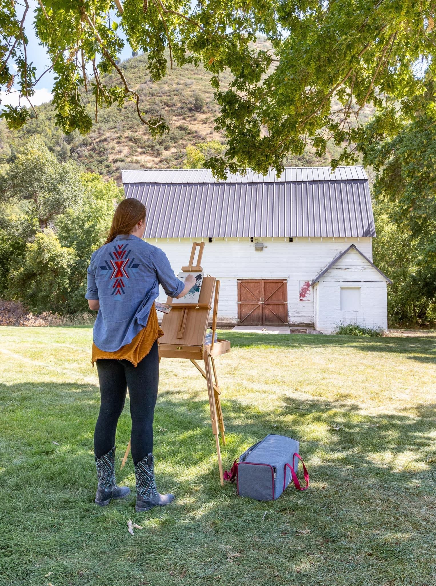 A woman is painting a barn in a field.