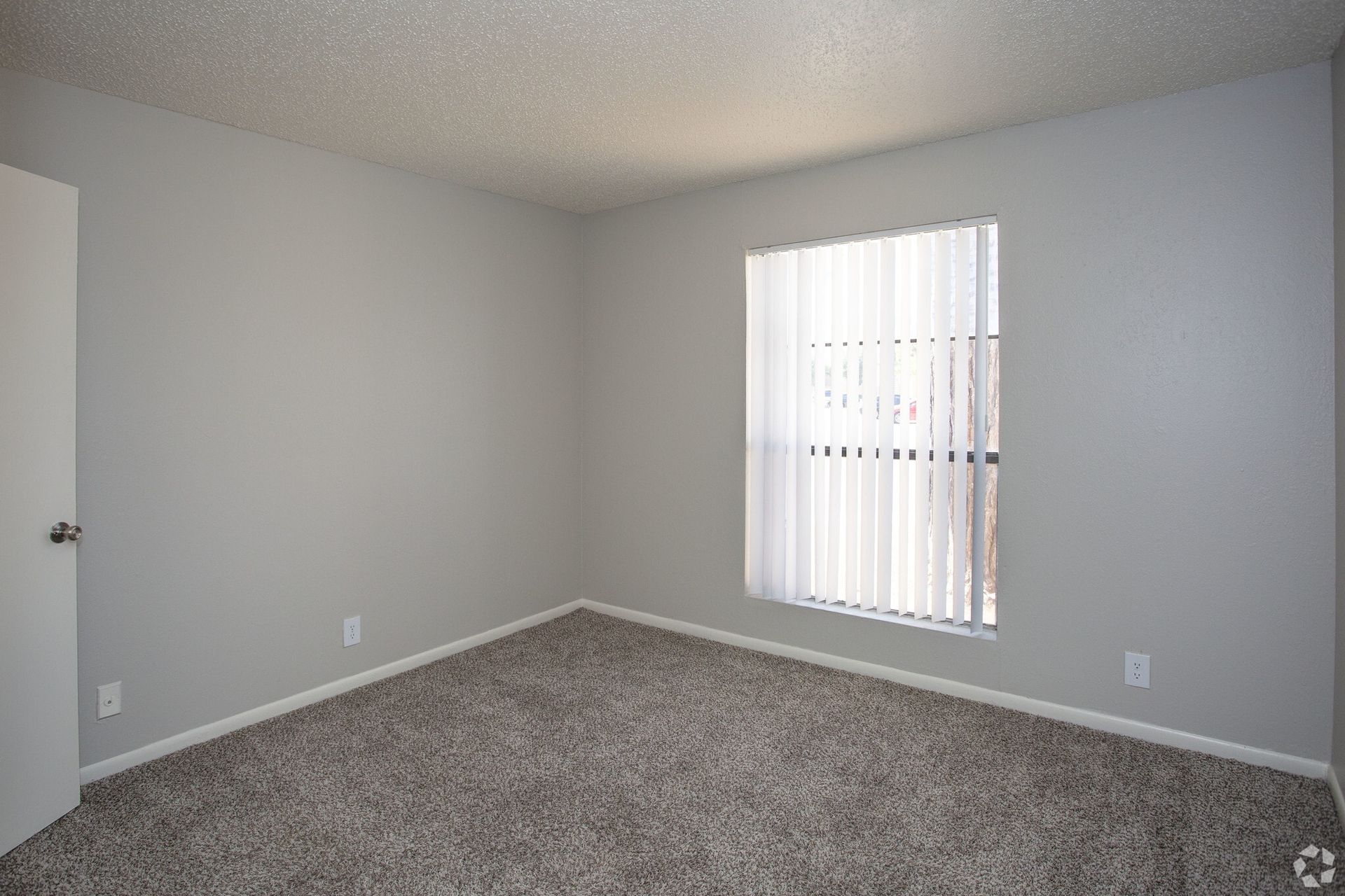Empty bedroom with neutral walls, carpet, and a window with vertical blinds at Woodview, offers apartments in Odessa, TX.