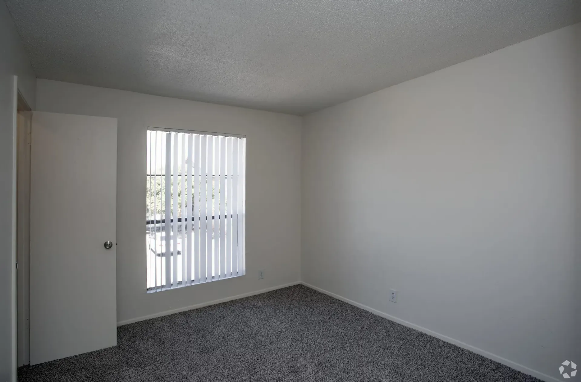 Empty white bedroom with a window covered by vertical blinds and carpeted floor at Woodview, offers apartments in Odessa, TX.