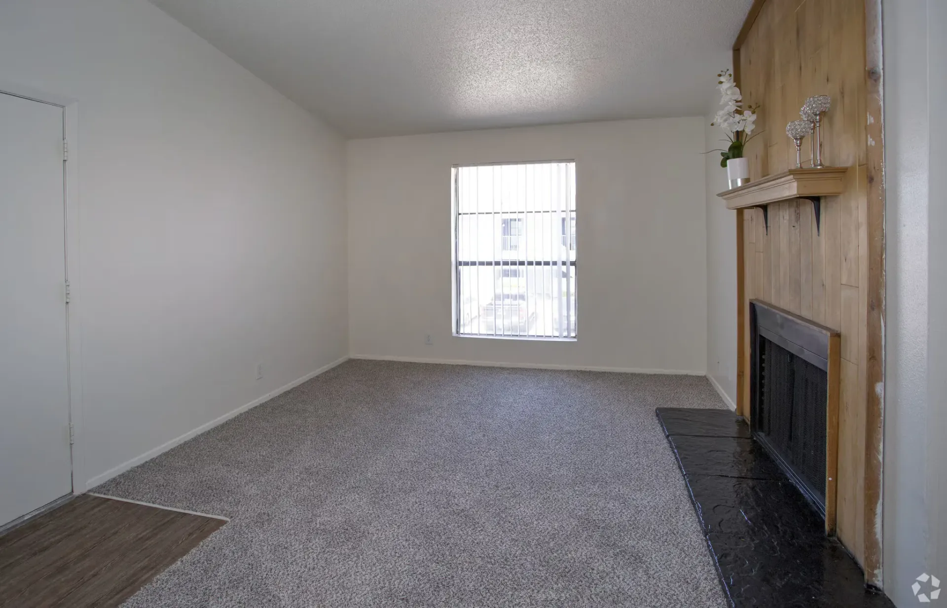 Empty living room with a large window, beige carpet, and a wood-paneled fireplace at Woodview, offers apartments in Odessa, TX.